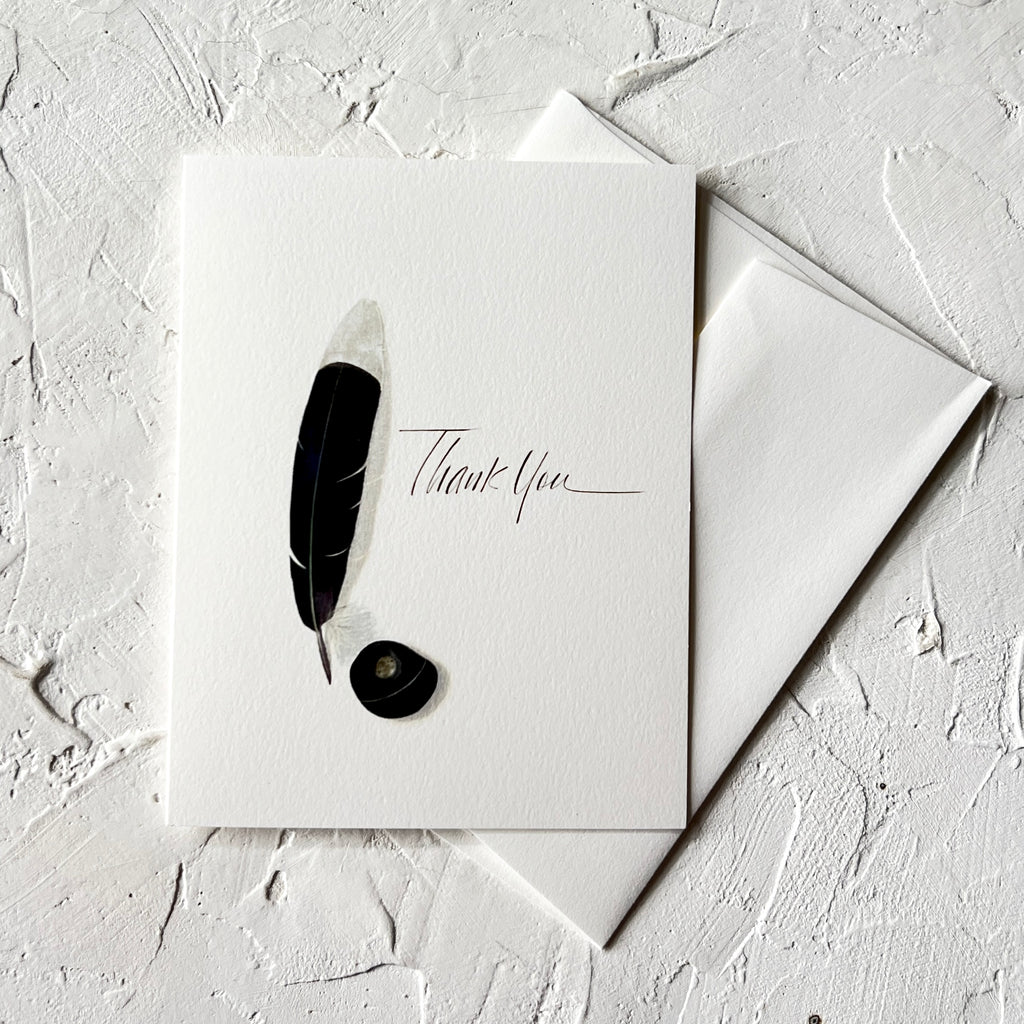 White notecards with black text saying, “Thank You”. Image of a black and white feather ink pen with a black inkwell. A white envelope is included.