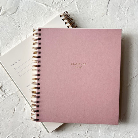 Notebook with dusty rose pink cover with gold foil text saying, “Gratitude Journal”. Gold coil binding on left side.