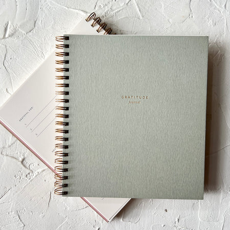 Notebook with sage green cover with gold foil text saying, “Gratitude Journal”. Gold coil binding on left side.