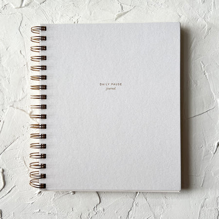 Notebook with white cover with gold foil text saying, “Daily Pause Journal”. Gold coil binding on left side.