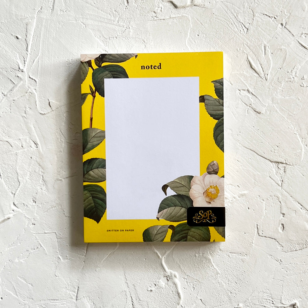 White notepad with yellow border and floral design with white flowers and green leaves. Black text saying, 