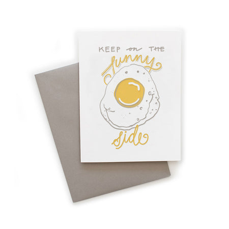 White card with gray and yellow text saying, “Keep On the Sunny Side”. Image of a sunny side up egg with a smiley face. A gray envelope is included.