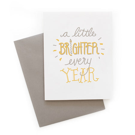 White card with gray and yellow text saying, “A Little Brighter Every Year”. Images of a gray and white birthday candle.  A gray envelope is included.