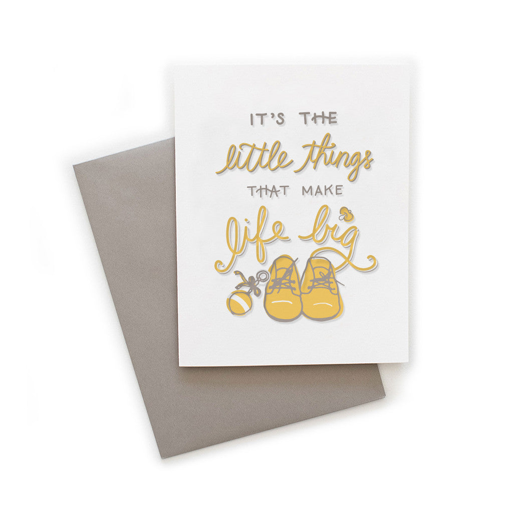 White card with gray and yellow text saying, “It’s the Little Things That Make Life Big”. Images of a pair of yellow baby shoes and a yellow baby rattle. A gray envelope is included.