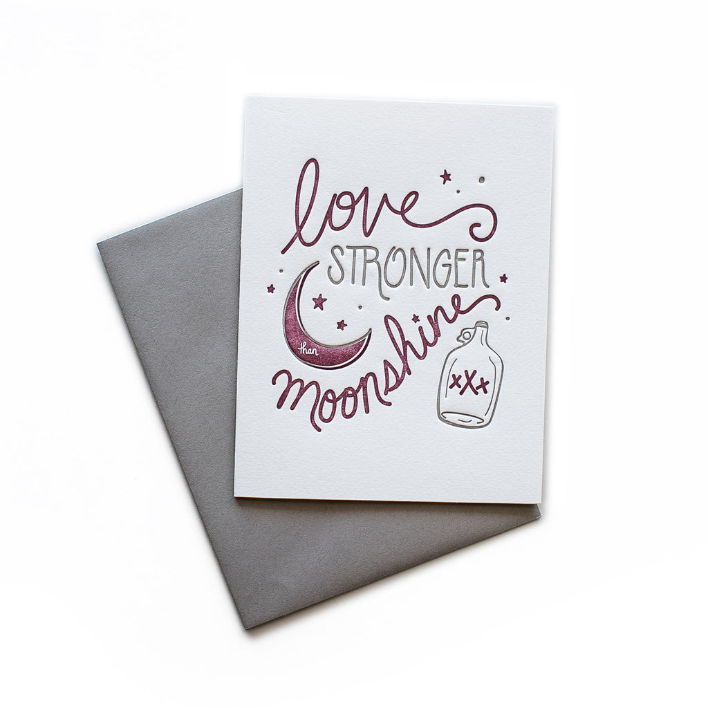 White card with gray and purple text saying, “Love is Stronger Than Moonshine”. Images of a purple moon and stars with a glass bottle of moonshine. A gray envelope is included.