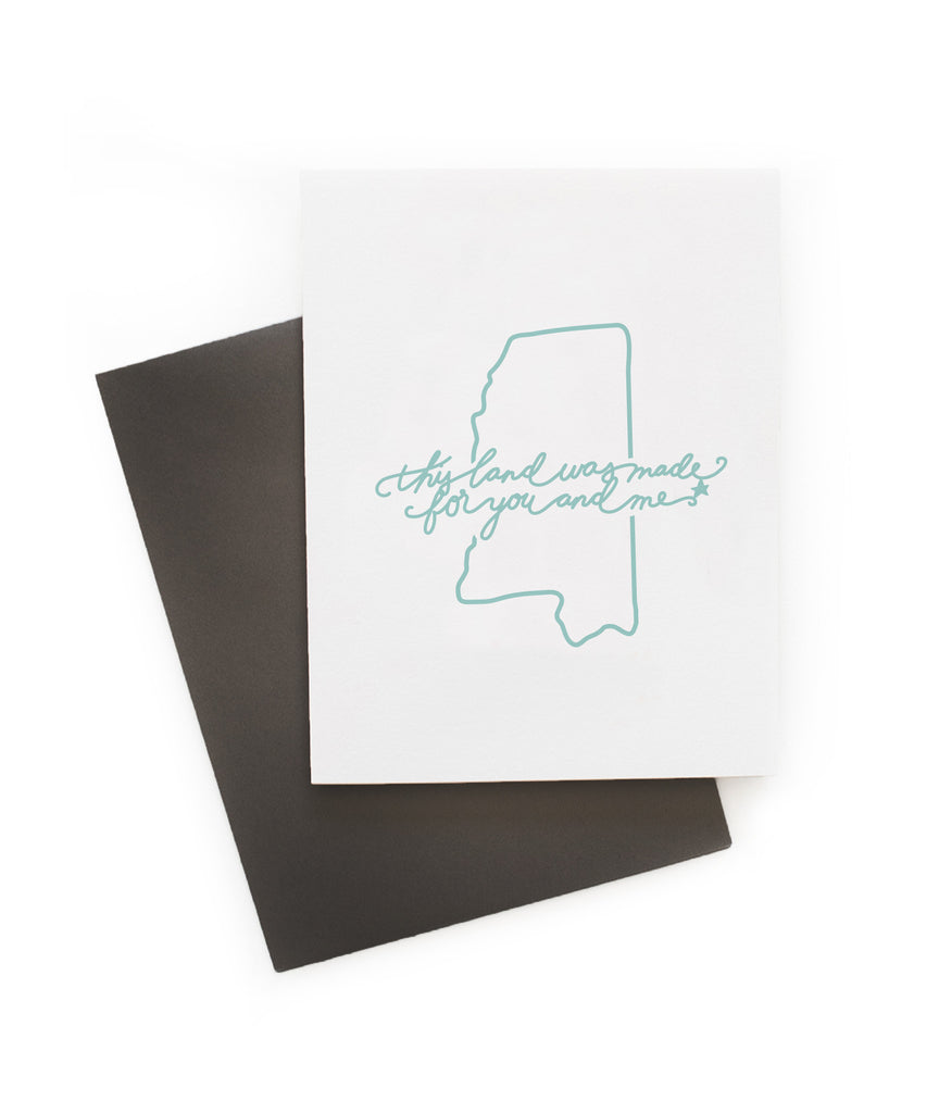 White card with teal text saying, “This Land is Made for You and Me”. Teal outline image of the state of Mississippi. A gray envelope is included.