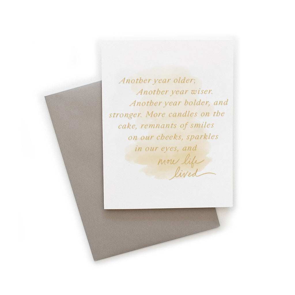 White card with yellow muted brushstroke background with yellow text saying, “Another year older, another year wiser. Another year bolder, and stronger. More candles on the cake, remnants of smiles on our cheeks, sparkles in our eyes, and more life lived”. A gray envelope is included.