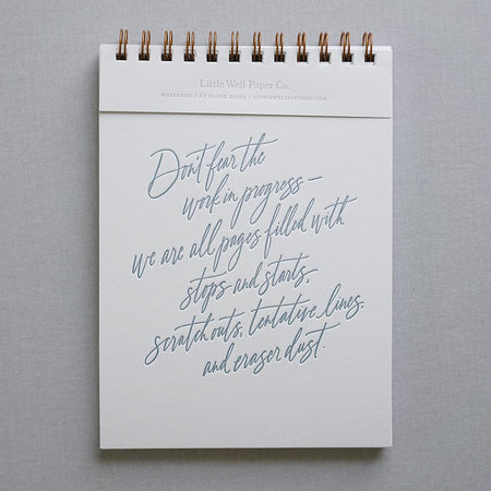 Notebook with white background and gray script text saying, “Don’t fear the work in progress- we are all pages filled with stops and starts, scratch outs, tentative lives and eraser dust”. Brass coil binding across the top.