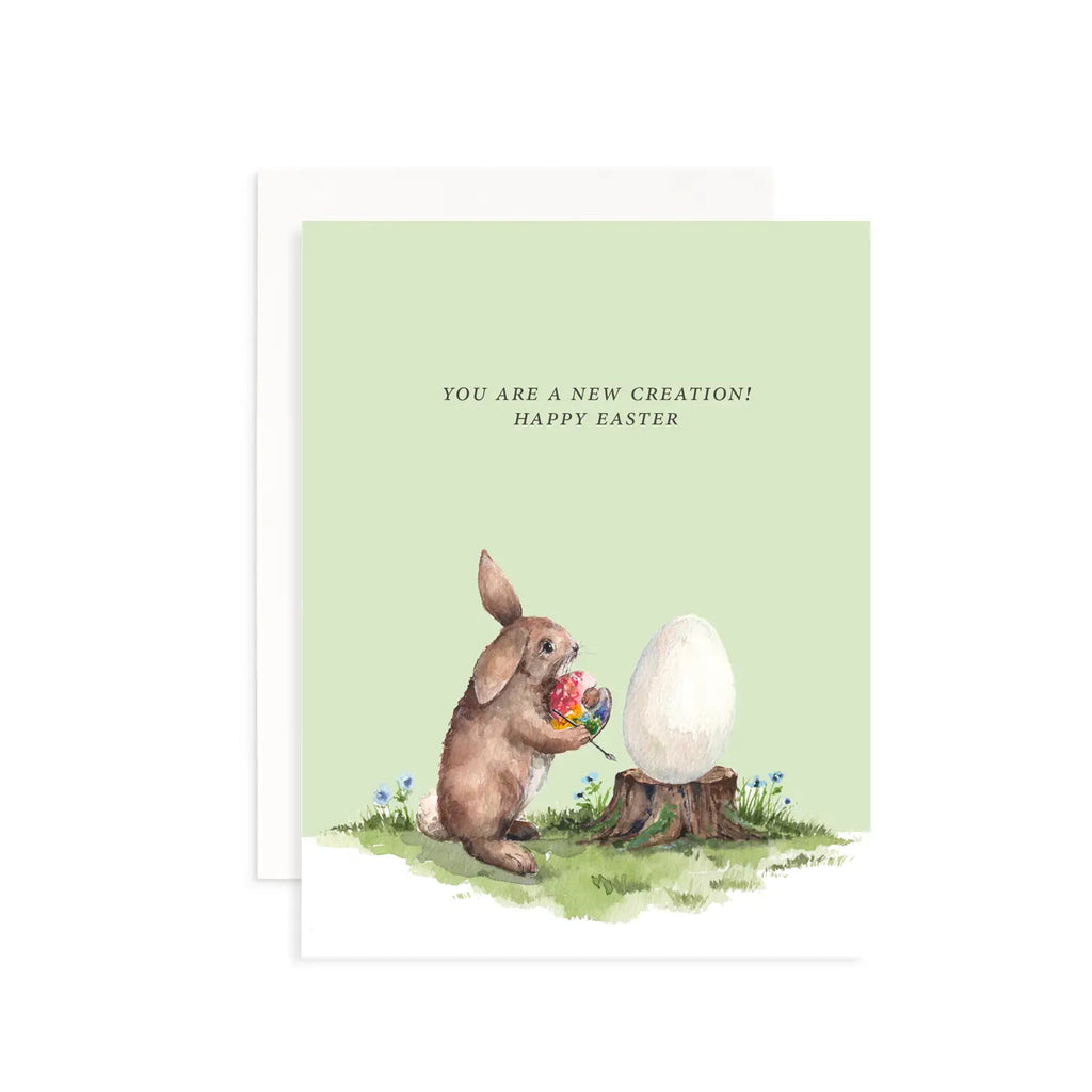 Mint green card with black text saying, “You Are A New Creation”. Image of a brown bunny holding a flower bouquet standing next to a white egg sitting on a brown tree stump. A white envelope is included.