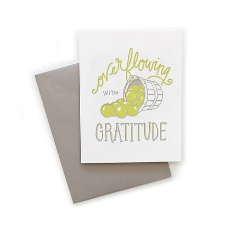 White card with gray and green text saying, “Overflowing with Gratitude”. Image of a bushel basket with green apples spilling out. A gray envelope is included.
