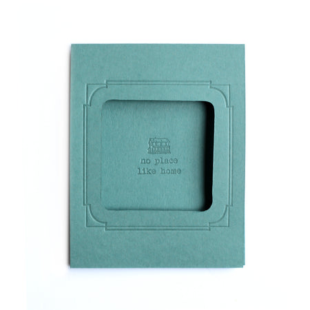Rectangle teal blue card with arched photo cut out in center with text saying, “No Place Like Home” with an embossed image of a house.  A matching envelope is included.