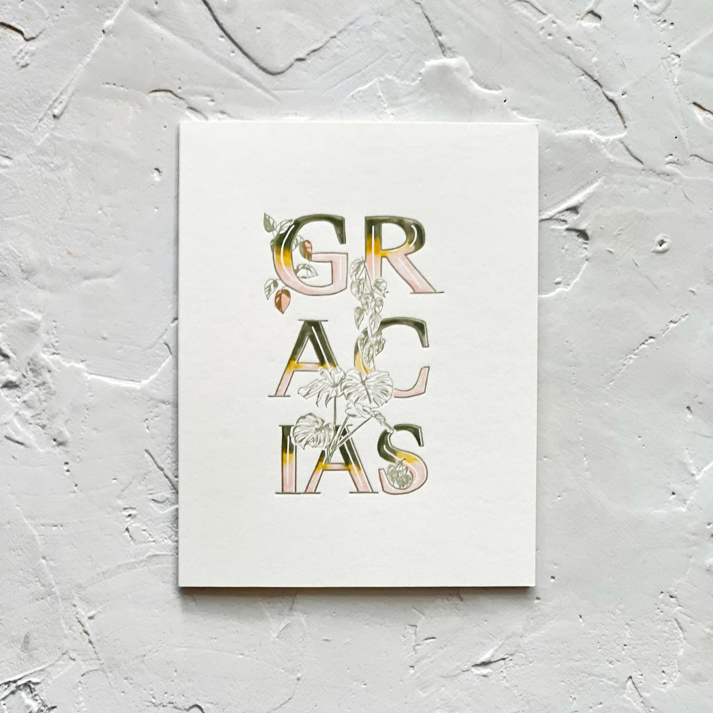 Ivory card with variegated text in green, yellow, white and tan saying, “Gracias”. Images of ivy vines entwined through the letters. An ivory envelope is included.