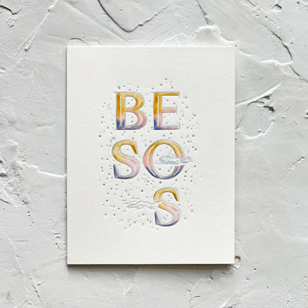 White card with rainbow text saying, “BESOS”. Images of clouds and stars. An envelope is included.