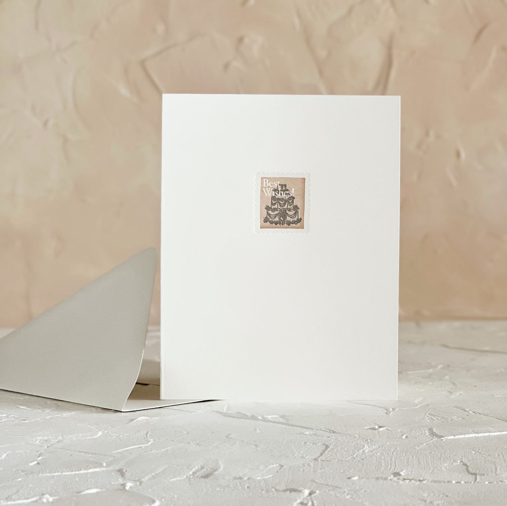 Ivory card with image of a postage stamp with a back 3 tier wedding cake and white text saying, “Best Wishes”. A gray envelope is included.