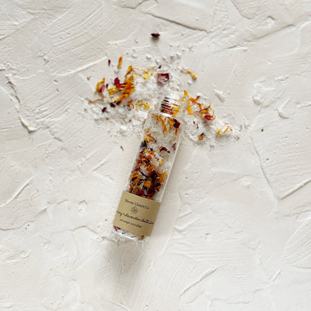 Small glass bottle with brown label saying, “Bloom Closet Co. Rosemary Lavender Bath Salts”. White salt mixed with brown, tan, orange and pink flecks of color.