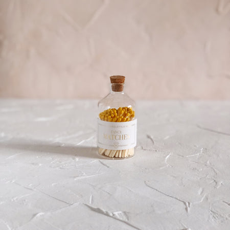 Small glass bottle with cork lid and white label with gold text saying, “Candlefolk Fancy Matches”. Filled with wooden matches with yellow tops.