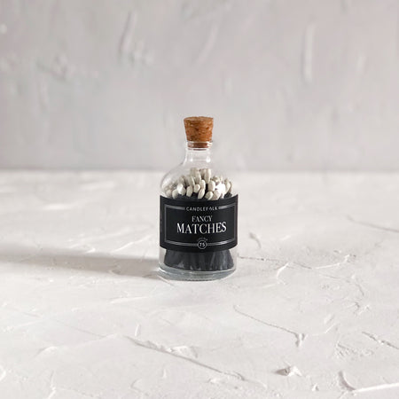 Small glass bottle with cork lid and black label with silver text saying, “Candlefolk Fancy Matches”. Filled with black wooden matches with white tops.