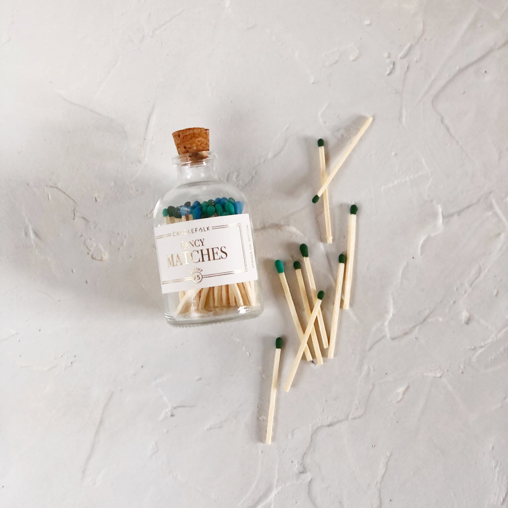 Small glass bottle with cork lid and white label with gold text saying, “Candlefolk Fancy Matches”. Filled with wooden matches with multicolored green, blue and black tops.