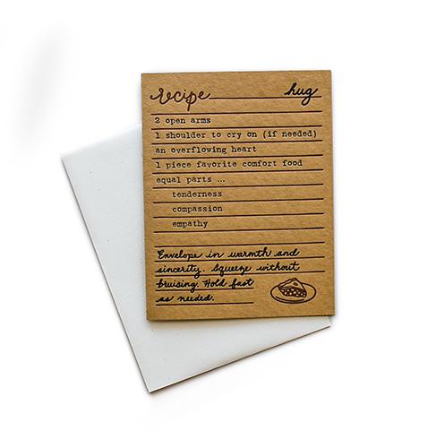 Brown card with black and brown text saying, “Recipe for Hug: 2 open arms; 1 shoulder to cry on; an overflowing heart; 1 piece of favorite comfort food; equal parts…tenderness; compassion; empathy. Envelope in warmth and sincerity. Squeeze without bruising. Hold fast as needed.” Image of slice of pie in bottom right corner. A white envelope is included.