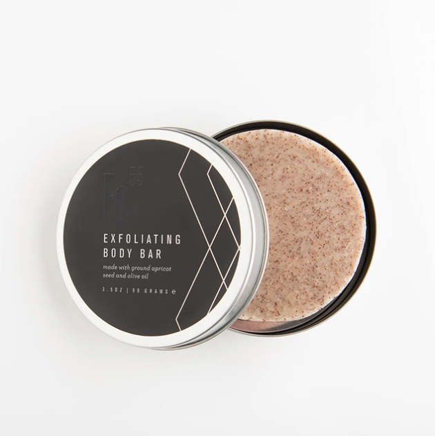 Packaged in a round black tin with silver and black lid. White text saying, “Exfoliating Body Bar”. Images of white geometric angles on side of lid. Body bar is light pink color.