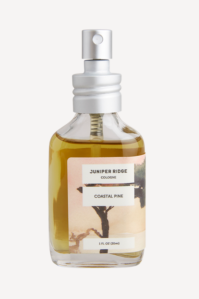 Small glass bottle with silver spritz topper. Label is pink with images of a coastal pine tree. Black text in an ivory box saying, “Juniper Bridge Cologne Coastal Pine”.