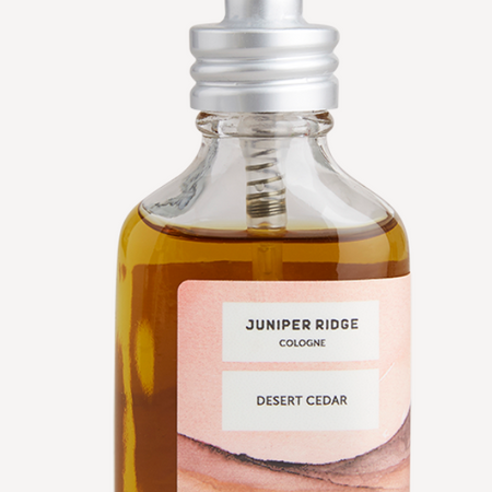 Small glass bottle with silver spritz topper. Label is pink with images of a muted desert. Black text in an ivory box saying, “Juniper Bridge Cologne Desert Cedar”.