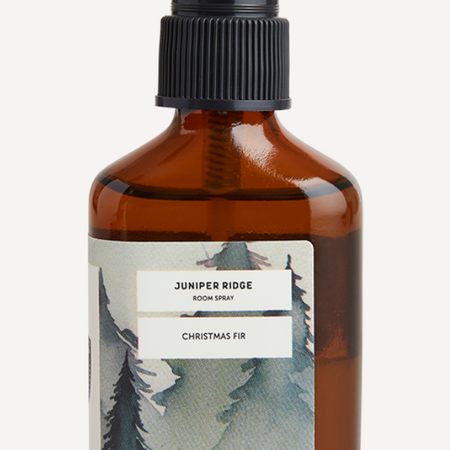 Small brown glass bottle with black spritz topper. Label is white with images of muted fir trees. Black text in an ivory box saying, “Juniper Bridge Room Spray Christmas Fir”.