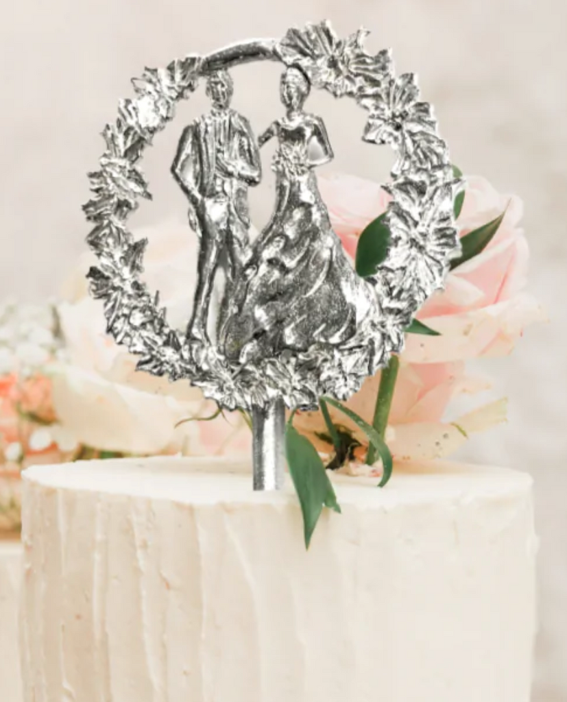 Silver pewter cake topper in the image of a bride and groom surrounded by a circle floral wreath.