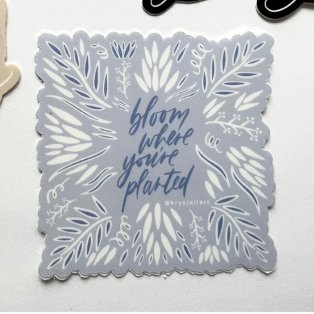 Light blue sticker with blue script text saying, “Bloom Where You’re Planted”.  Images of blue and white leaves.