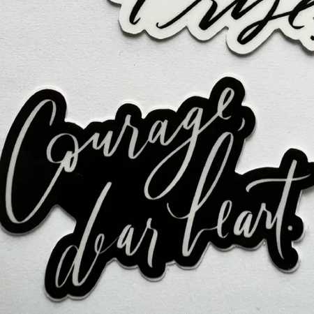 Black sticker with white script text saying, “Courage, Dear Heart”.