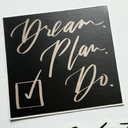 Black sticker with white script text saying, “Dream. Plan. Do.” Image of a square with a checkmark in it.