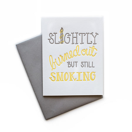 White card with gray and yellow text saying, “Slightly Burned Out But Still Smoking”. Images of a yellow birthday candle. A gray envelope is included.