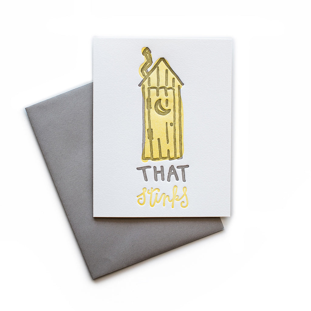 White card with gray and yellow text saying, “That Stinks”. Image of a yellow wooden outhouse with moon cutout on door. A gray envelope is included.