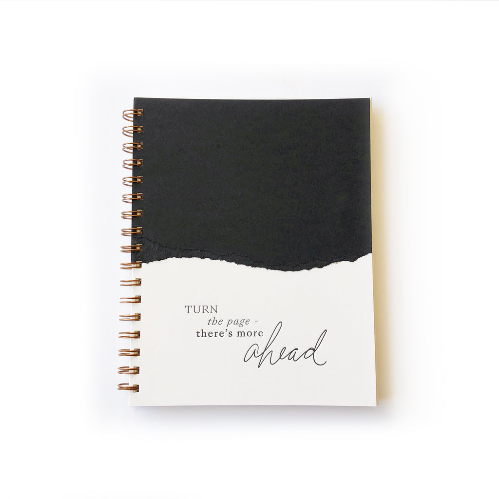 Notebook with ivory and black curved torn edging where the two colors meet on cover with black text saying, “Turn the Page There’s More Ahead”. Brass coil binding on left side.