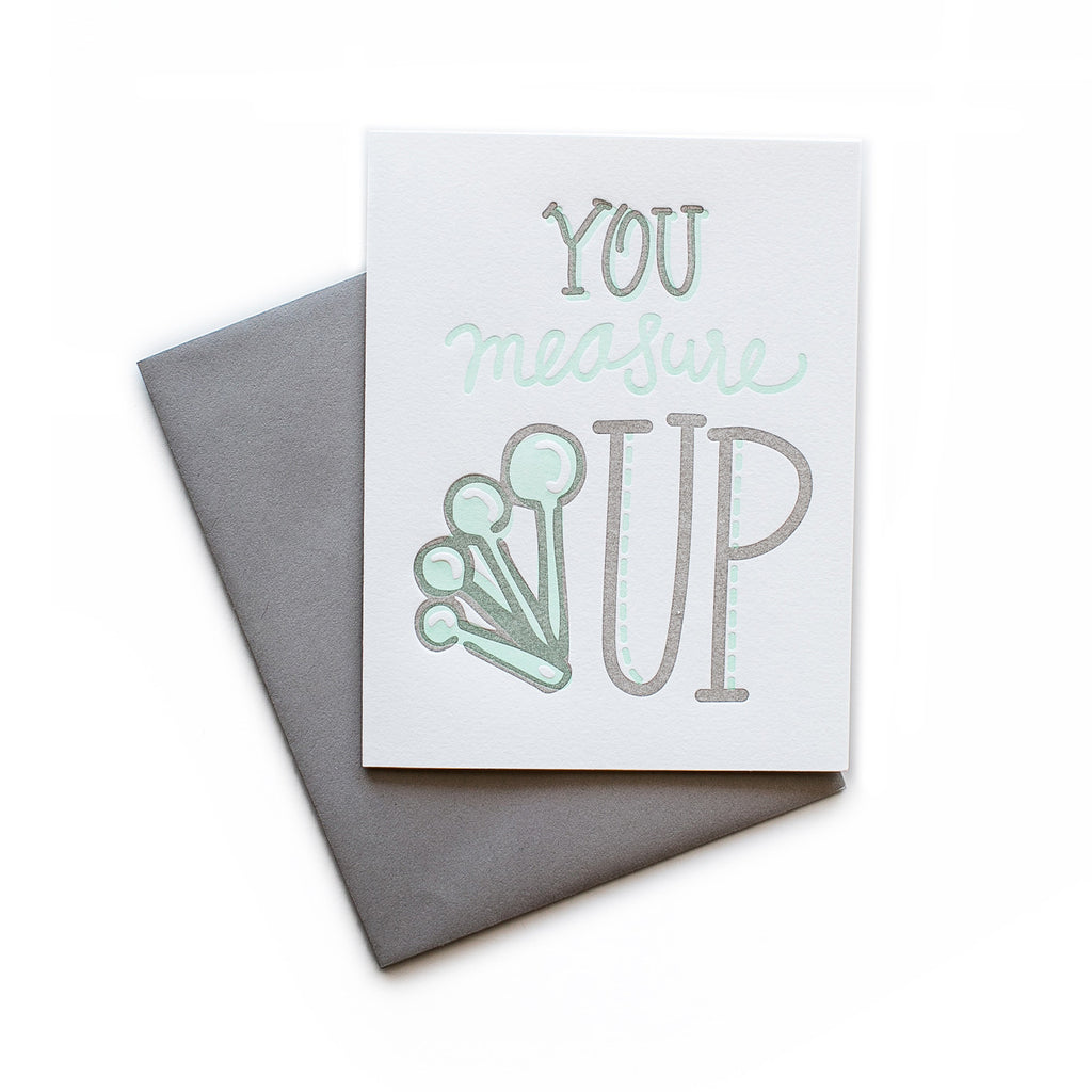 White card with gray and teal text saying, “You Measure Up”. Image of a set of teal measuring spoons. A gray envelope is included.