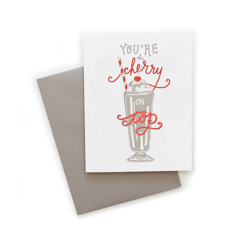 White card with gray and red text saying, “You’re the Cherry on Top”. Image of a gray ice cream float with a red cherry on top and a white and red straw. A gray envelope is included.
