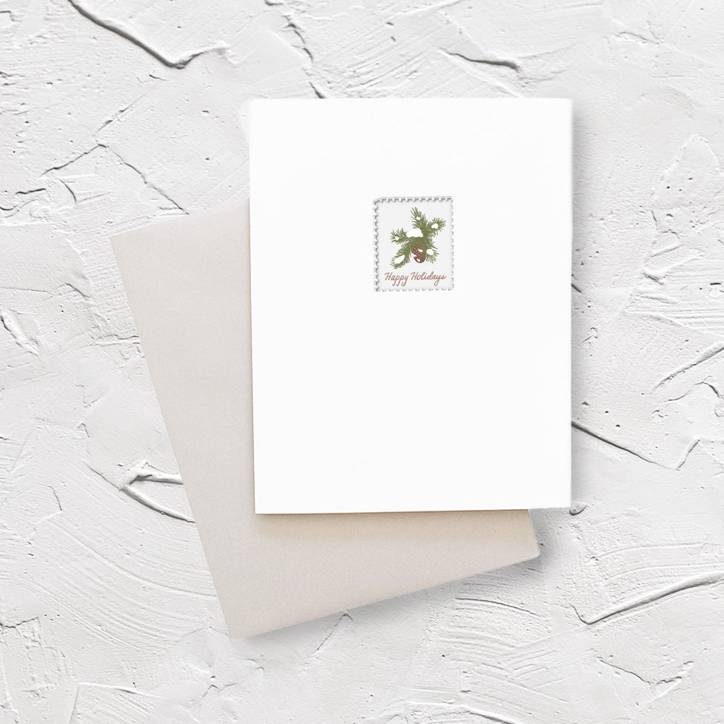 White card with image of a postage stamp with white background and evergreen branches with brown pinecone. Red text saying, “Happy Holidays”. A gray envelope is included.