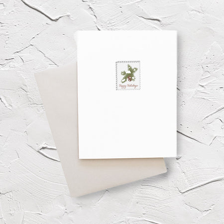 White card with image of a postage stamp with white background and evergreen branches with brown pinecone. Red text saying, “Happy Holidays”. A gray envelope is included.