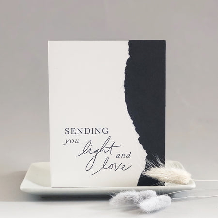 White and blue card with curved torn edging where the two colors meet. Blue text saying, “Sending You Light and Love”. A white envelope is included.