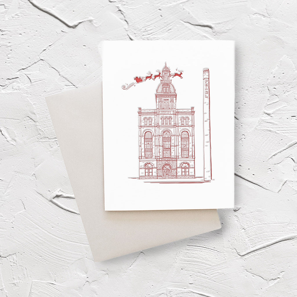 Ivory card with red ink. Image of a building from the Pearl District in San Antonio with Santa and his sleigh flying over it. A gray envelope is included.