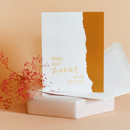 White and orange card with curved torn edging where the two colors meet and orange text saying, “You Will Never Walk Alone”. A white envelope is included.