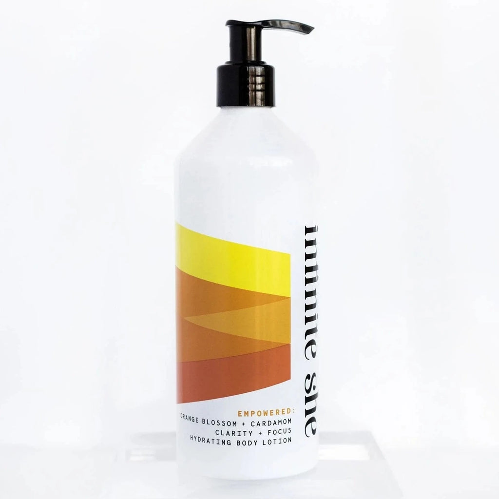White bottle with black pump top and black text saying, “Infinite She Empowered Hydrating Body Lotion”. Images of yellow, brown, and orange triangle design.