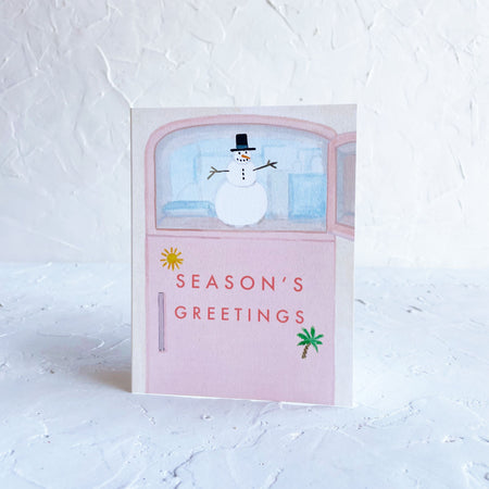 White card with red text saying, “Season’s Greetings”. Image of a pink refrigerator with a white snowman and ice cubes in the freezer. Image of a yellow sunshine and green palm tree on front of refrigerator door. A white envelope is included.